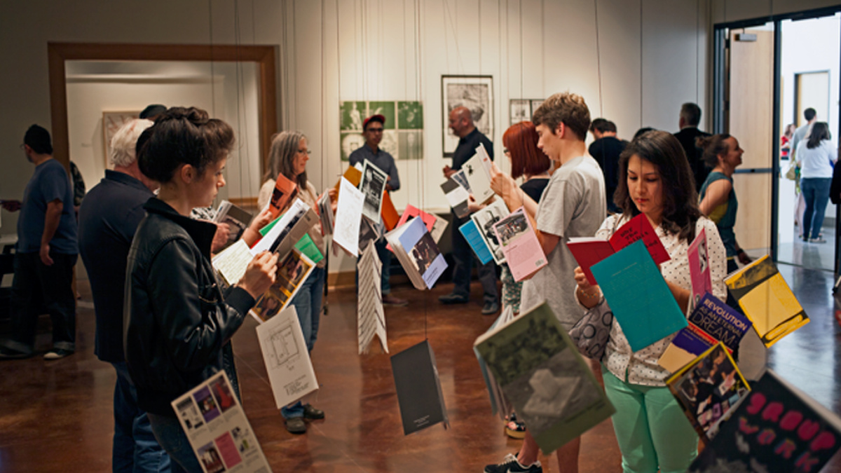 A photograph from an exhibition which features several books hanging from the ceiling. People hold and read the books.