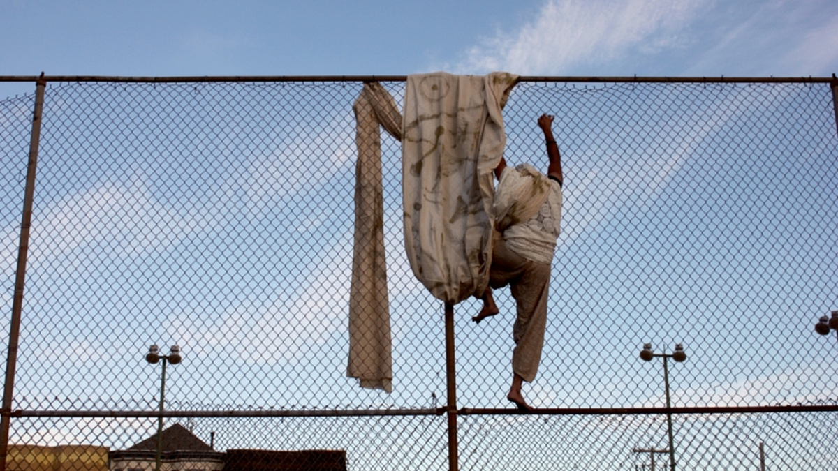 A photograph of a person climing a wire fence. A large piece of fabric is draped over the fence.