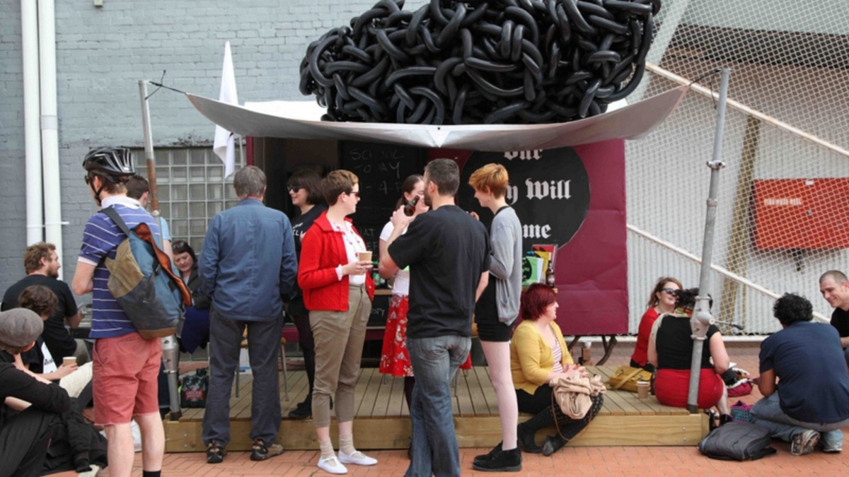 A group of people stand and sit on an outdoor patio, under a large sculpture of a knot of chains.