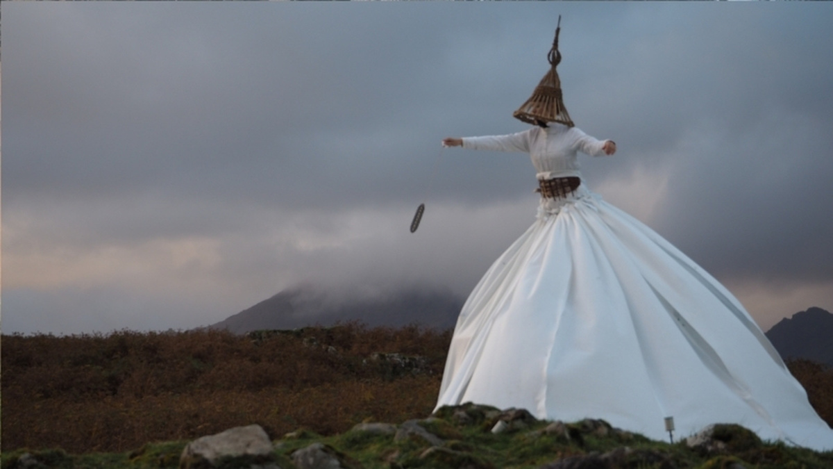 Artist Hanna Tuulikki wears a white dress with an exaggerated skirt standing in a misty, mountainous landscape.