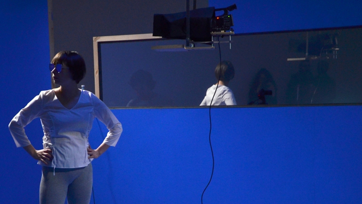A person stands wearing a long sleeved white tshirt, with their hands on their hips in front of a blue background.