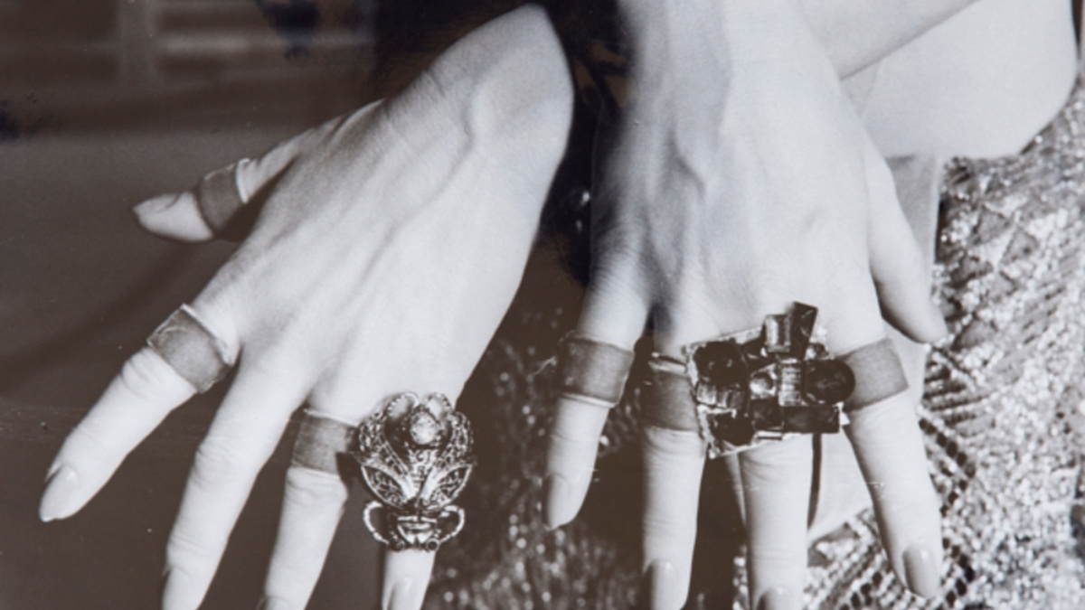 A black and white photograph of a pair of hands adorned with large jewel rings and ribbons around each finger.