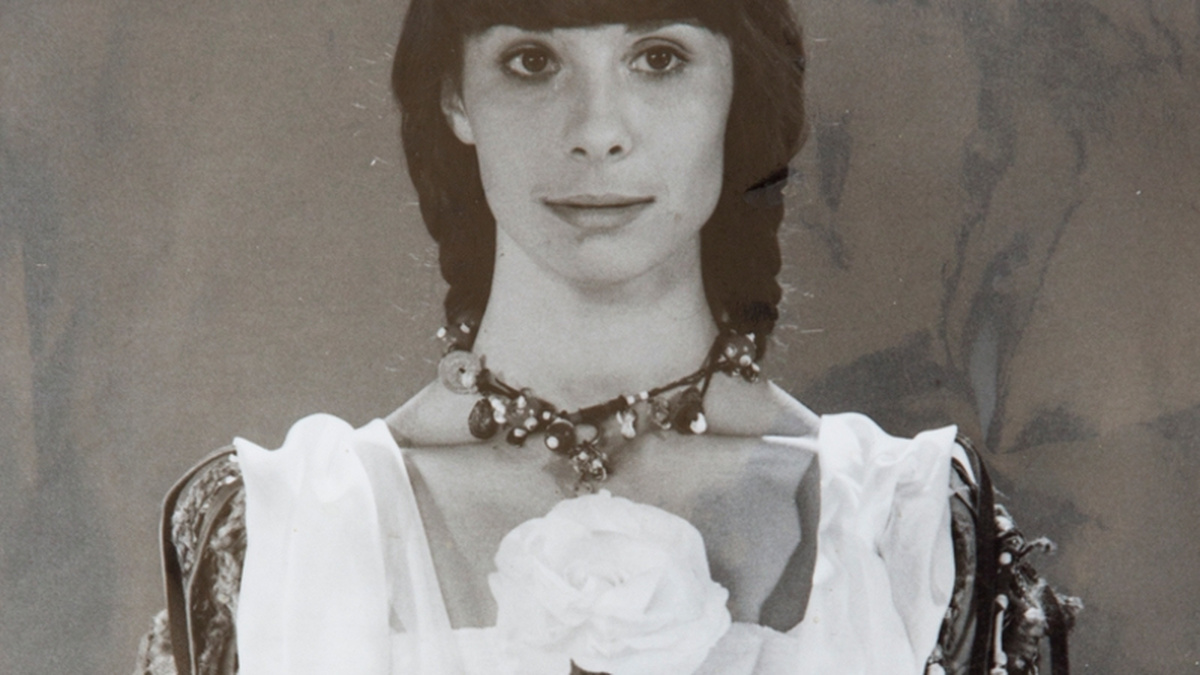 A photograph of a woman from the torso up, she is wearing a jeweled necklace and a dress, holding a large white rose.
