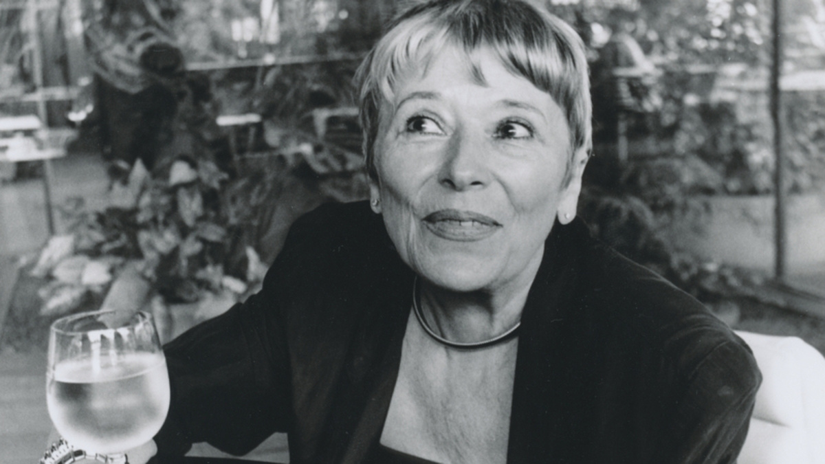 An black and white photograph of Ester Krumbachová, she has cropped hair and a dark blazer, holding a glass of wine.