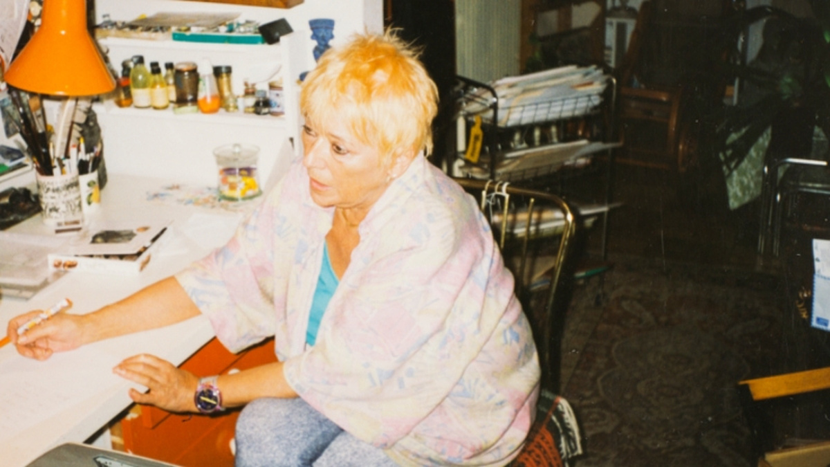A photo of Ester Krumbachová, she has cropped blonde hair and sits at a desk with a pen in her hand.