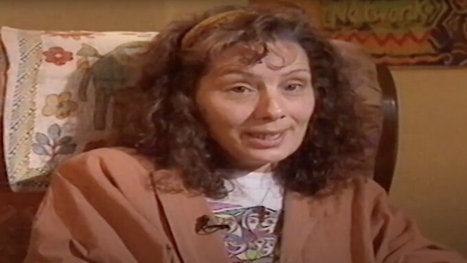 A woman with brown curly hair is sitting in an armchair, talking to the camera.