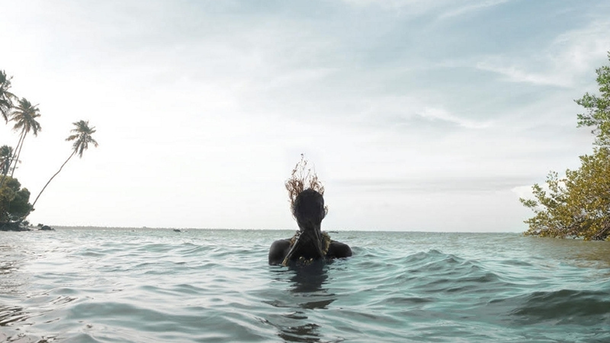 A photograph of a person emerging from the water, there are blue skies and tropical islands in the background.