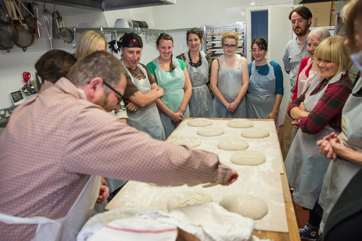 Twelve people stand round a kitchen worktop making bread dough.