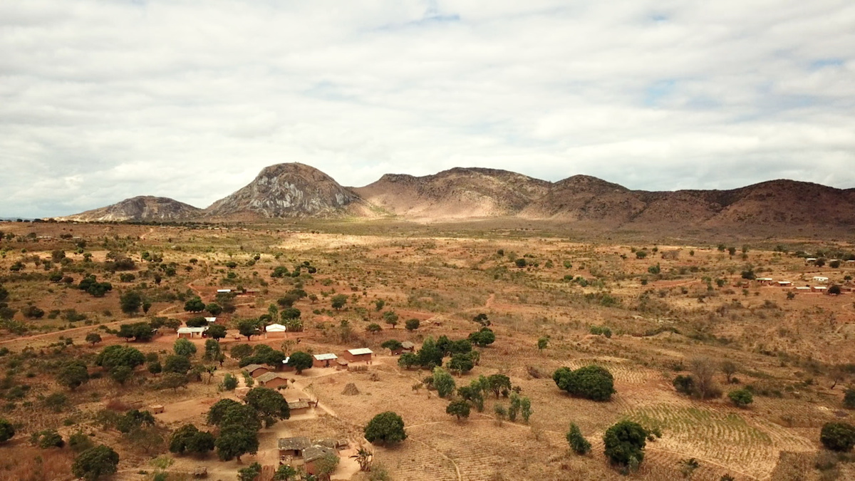 Several small houses with white roofs in a vast expanse of dry land. Above a distant mountain range, a cloudy sky.