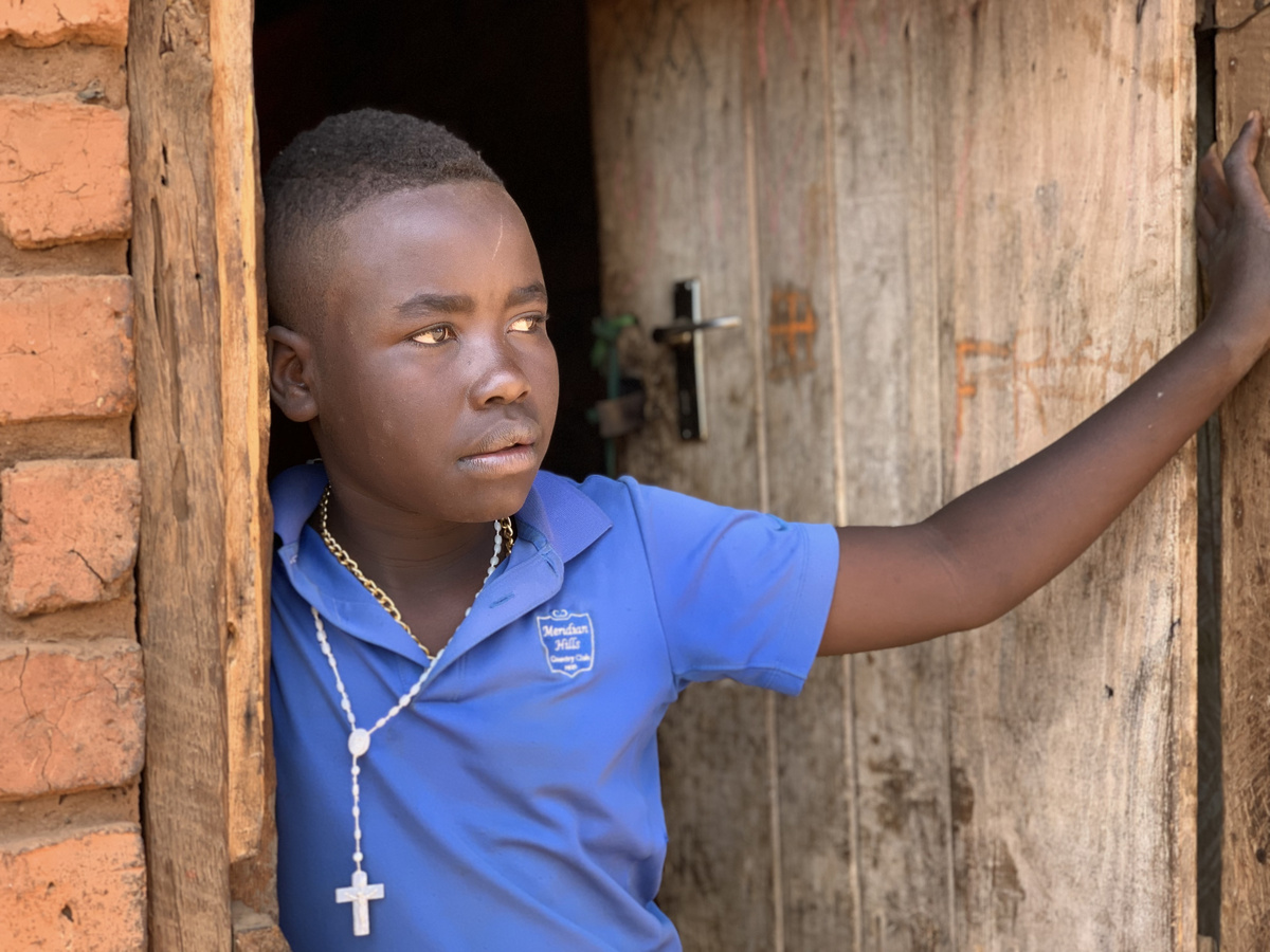 A child leans against a door with a pensive expression, in a blue polo shirt with a school crest and a rosary necklace.