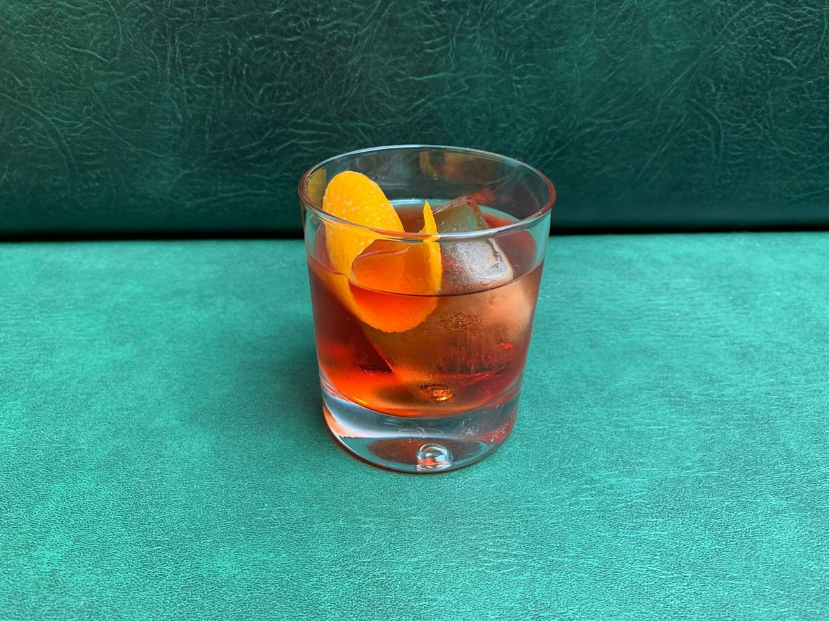 A bright orange cocktail with a slice of orange in a short glass, with a turquoise background.