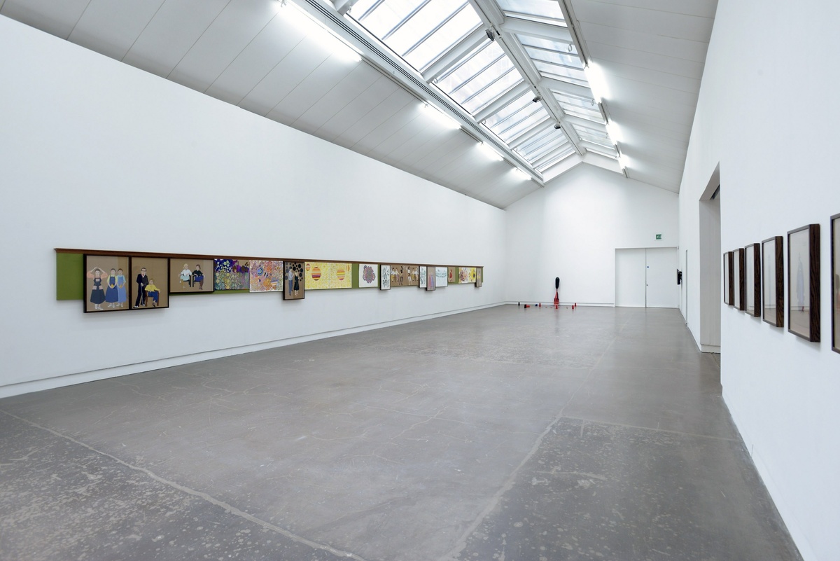 A long gallery space with white walls and grey flooring. Individual artworks hang on the left and right walls.