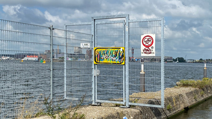 A view through a mesh fence of a body of water and an urban riverscape. There are panels with graffiti on the fencing.
