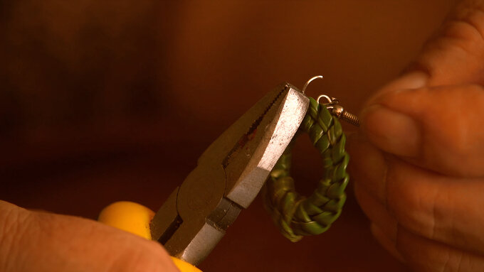 A close up of two hands holding metal pliers and an earring hook, fastening the hook to a small ring of knotted grass.