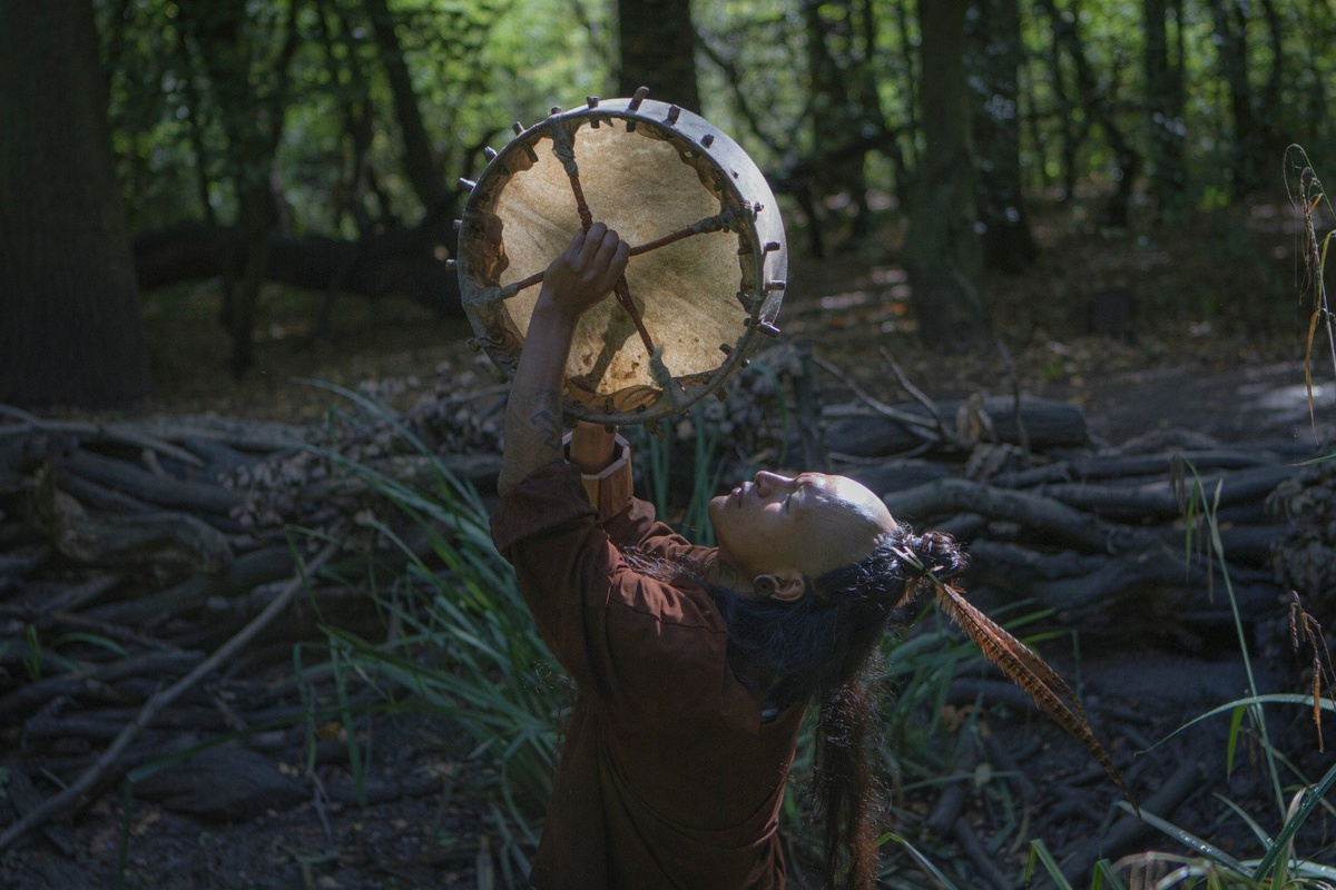 A photo of Ruben Vucubcame standing in a forest, wearing a long feather in their hair and holding a drum up to the sky.