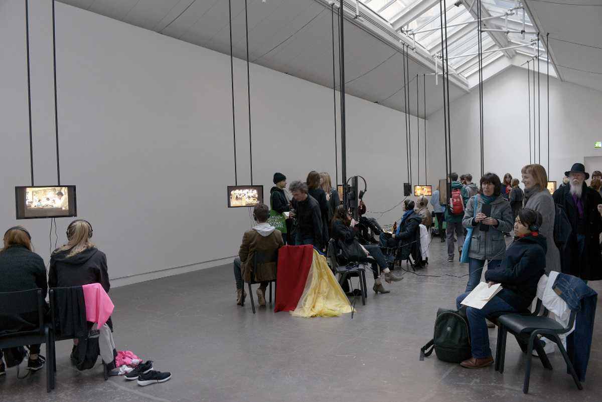 A photograph of the CCA gallery space, there are screens hanging from the ceiling and groups of people crowd around them