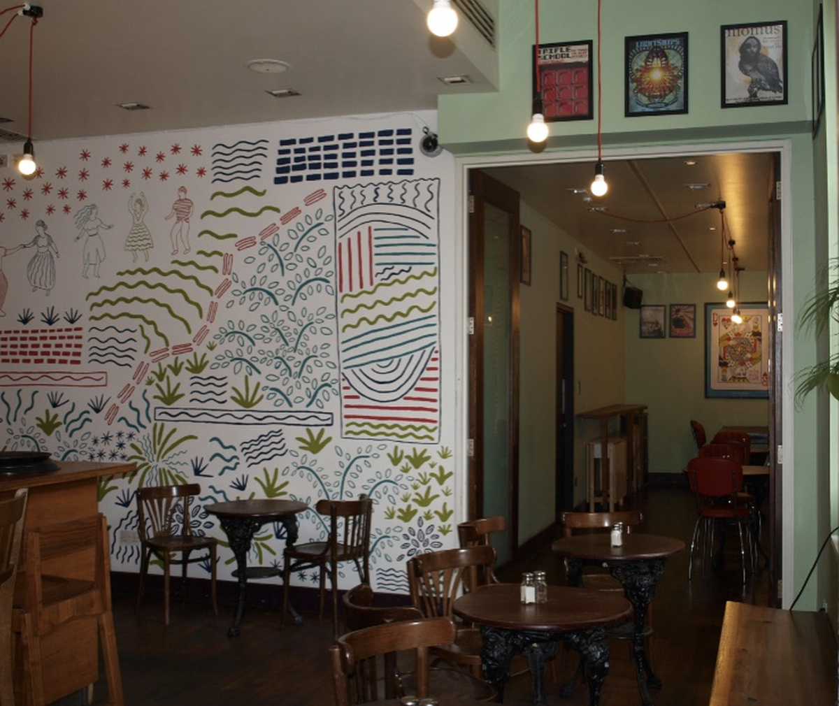 A photograph of the terrace bar interior, featuring wooden seats and round wooden tables and a large wall mural.