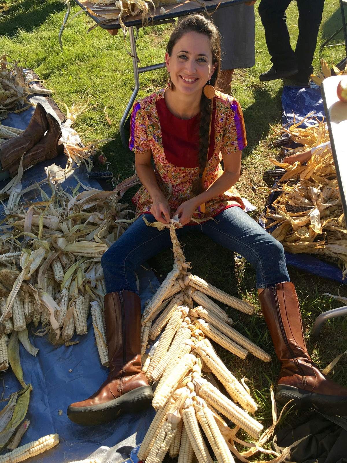 A Mohawk/Mi'kmaq woman with dark plaited hair sits on grass surrounded by corn husks, hand-braiding harvested corn