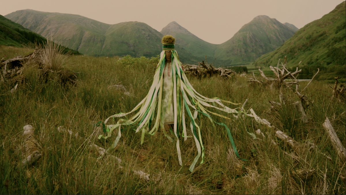 A costumed folkloric femme figure stands in the Glen Etive Forest’s rewilding project in embroidered ankle-length blouse