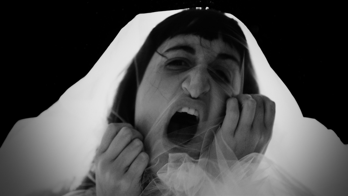 A closeup black and white image of a person's face under a clear veil, they are clawing at it, looking angry, desperate.