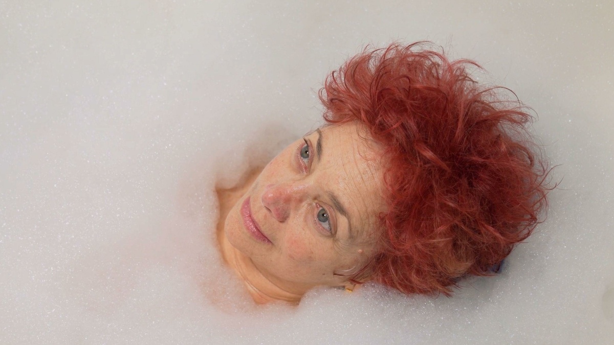 A white nonbinary person with red hair lying in the bath looking up at the ceiling. They are surrounded by bubbles.
