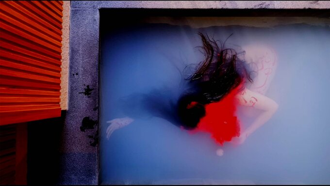 A person with long black hair is submerged in milky water in a pool. Their face is covered by a thick, bright red liquid
