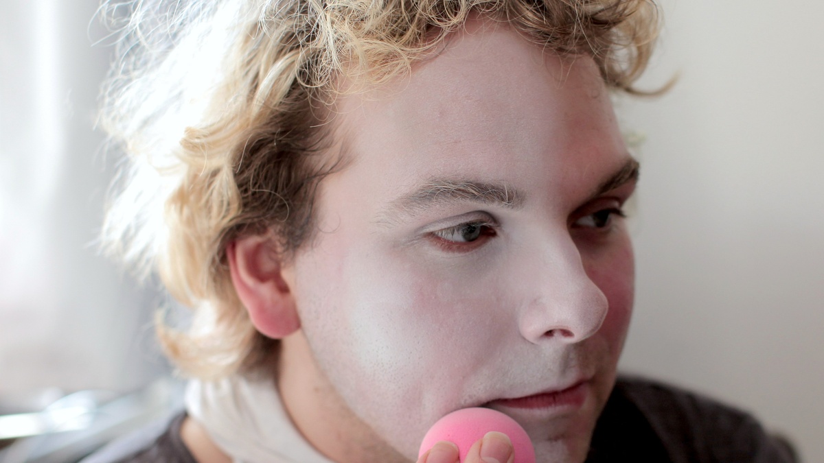 A close up of the face of a person as they apply white make up to their face with a pink make up sponge.