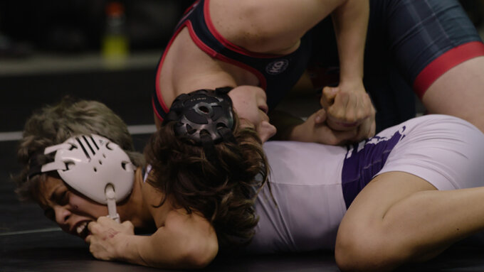 A close up of two white people wrestling. They wear head gear and sports tunics. The image feels dynamic and active.