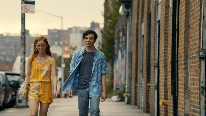 Two young people walking down a city street: a white woman in a yellow jumpsuit and the other is a white man in jeans.