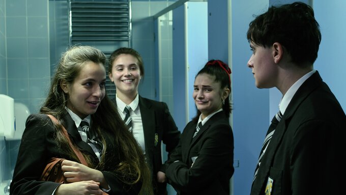 Three school girls stare at a boy with vicious smiles on their faces in a school bathroom. The boy looks worried.