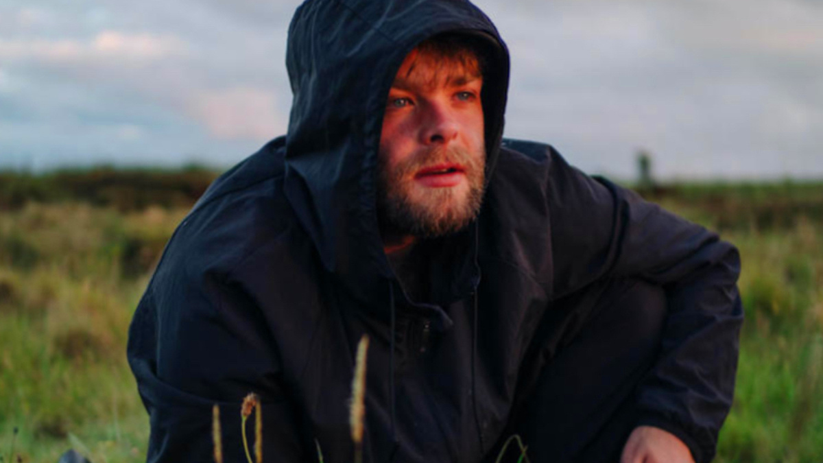 A person with a beard is crouched down in long grass and looking into the distance, wearing a dark anorak.