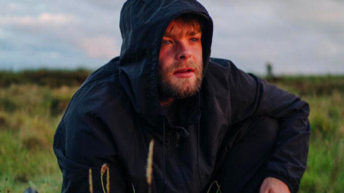 A person with a beard is crouched down in long grass and looking into the distance, wearing a dark anorak.