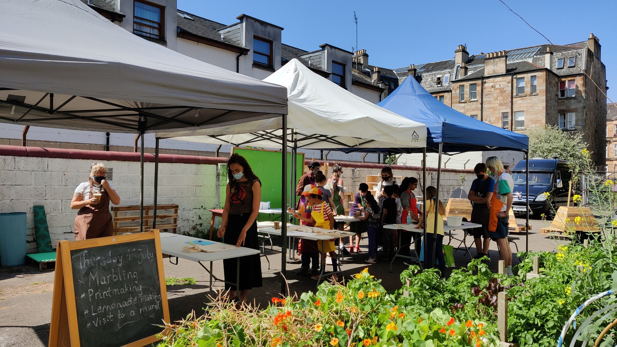 A photograph of a sunny outdoor square with market stalls and lush green foliage.