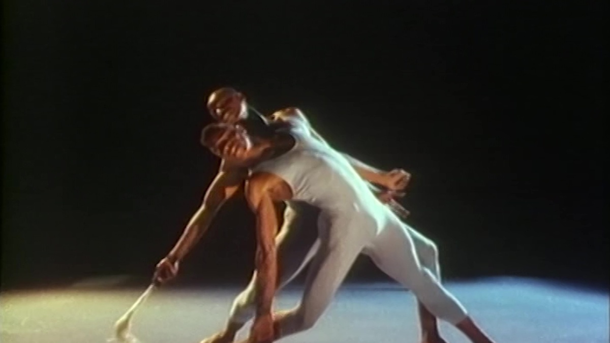 Two figures in grey leotards pose athletically in a dark room, illuminated by spotlight.