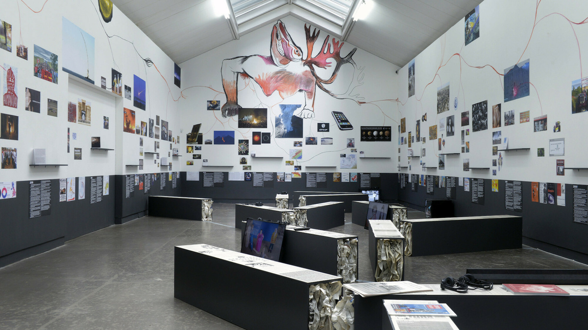 A gallery space with benches and lots of colourful images and text on three white and grey walls.