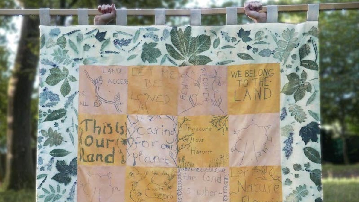 A quilted banner hanging from a wooden pole, squares of pink and yellow fabric inscribed with messages and images.