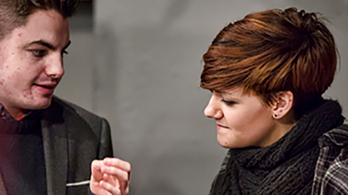 An image of a young person with short red hair and eye-liner in conversation with another person in a grey suit jacket.