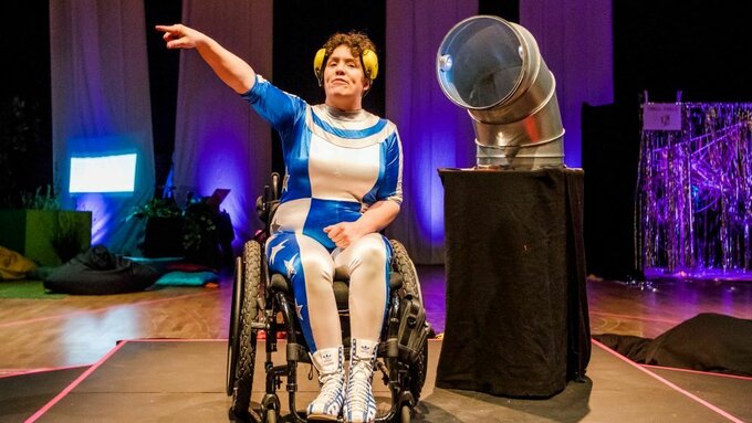 Jess, a woman with curly brown hair, is wearing a blue and white jumpsuit. She is sitting onstage in her wheelchair.