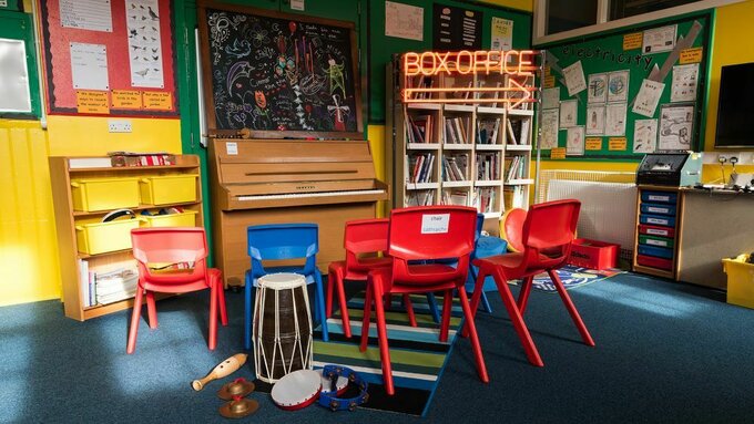 A brightly coloured school classroom with blue and red chairs, yellow walls and a neon sign that says "Box Office"