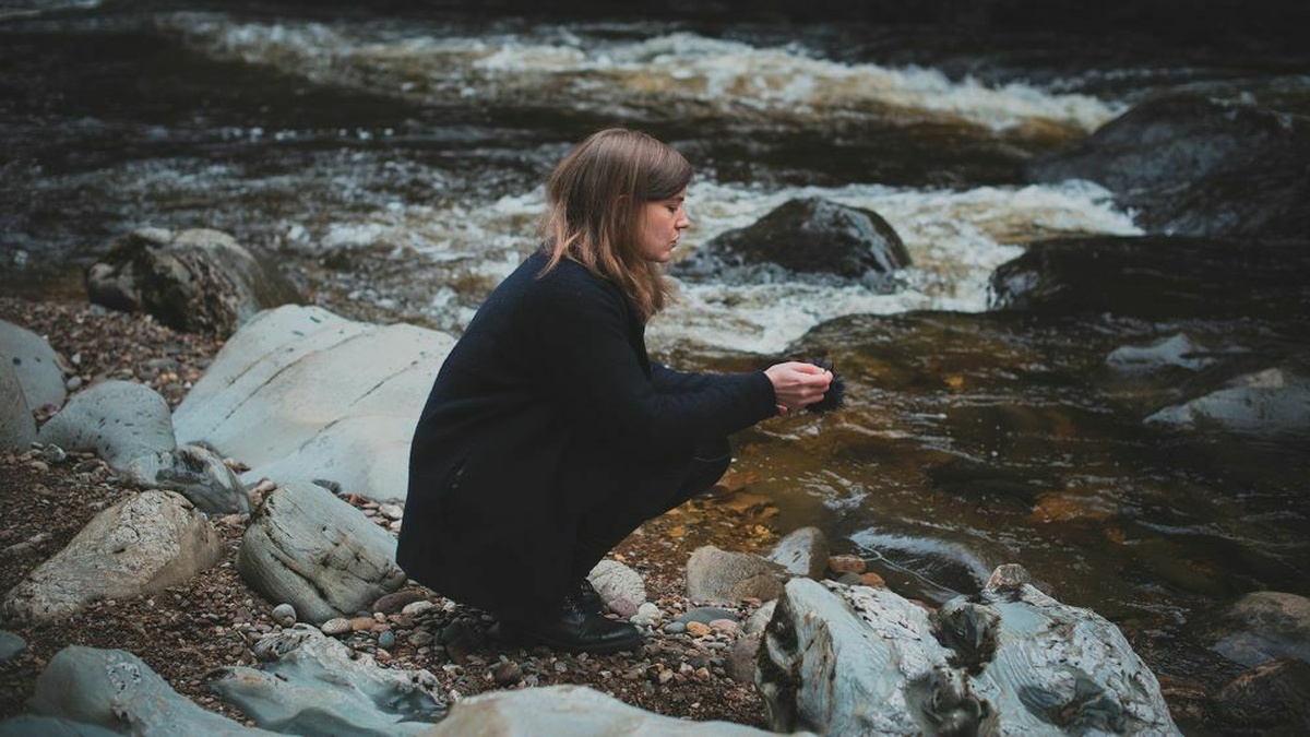 A woman crouches pensively on the rocky banks of a river, wearing a long dark coat, boots and is staring into the water.