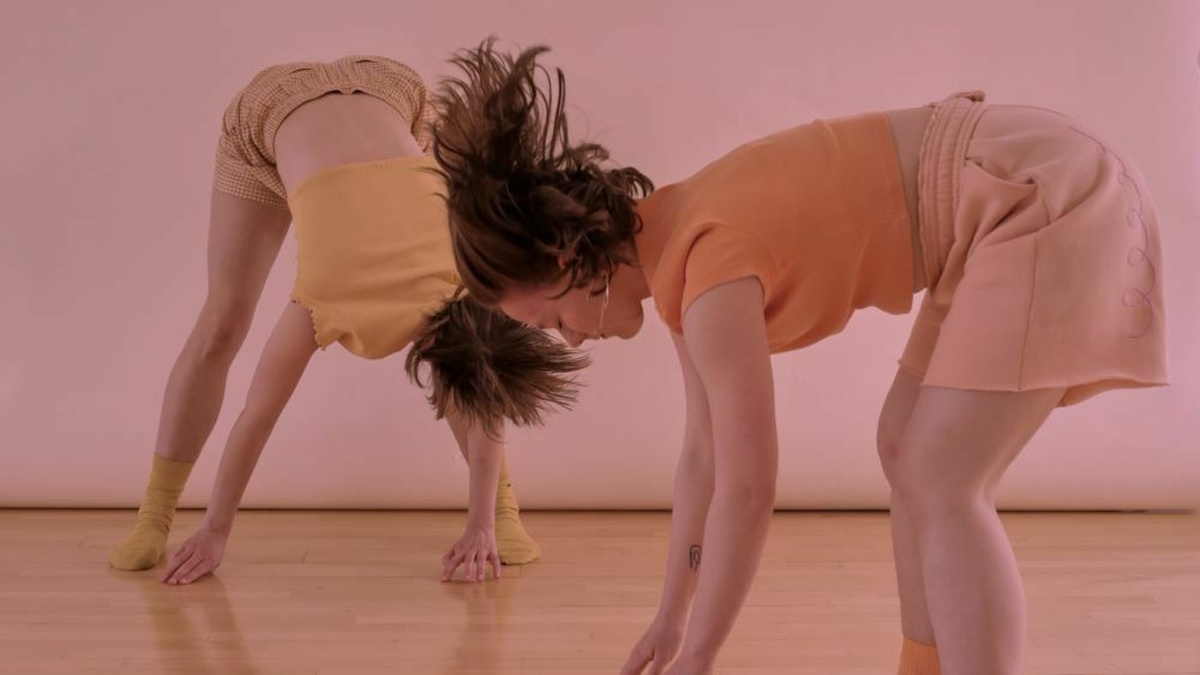 Two dancers wear comfortable pastel orange, yellow and pink clothes. They are both in the middle of touching the ground.