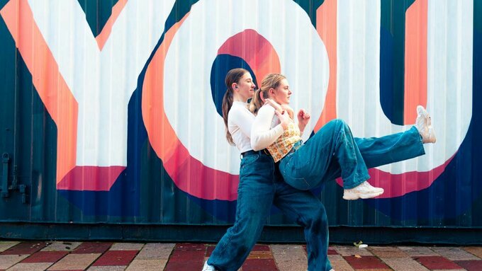 Two woman dance in front of a blue metal shipping container with the word "You" painted on it in white and pink letters.