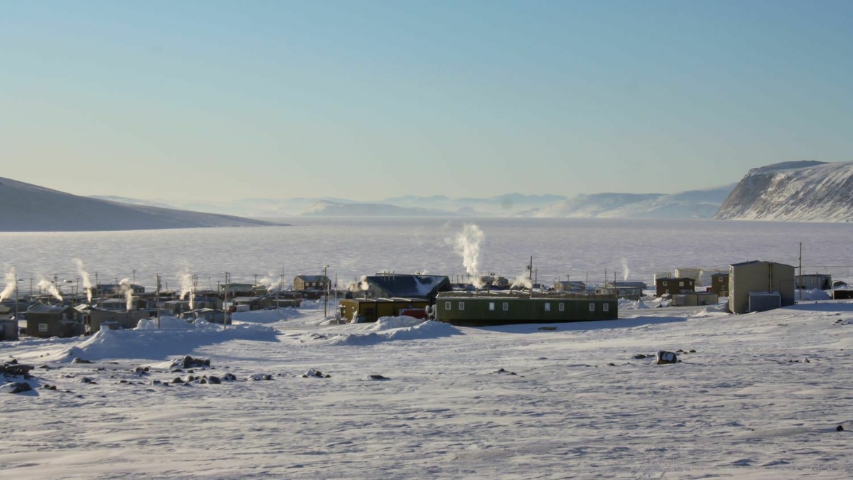 A photograph of a snowy landscape with a group of buildings in the centre. There are mountains in the background.