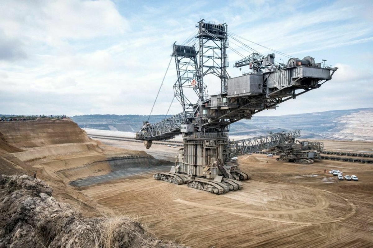 A giant grey machine consisting of cranes and drills stands in a desert landscape, having made markings in the sand.