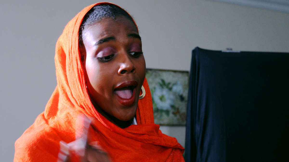 Black woman with bright orange shawl wrapped around her head sings, looking away from the camera.