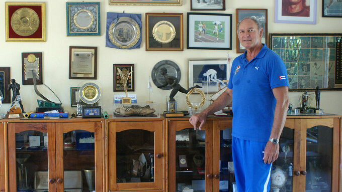 A man wearing a blue top and trousers stands leaning against a display cabinet containing many trophies and awards.