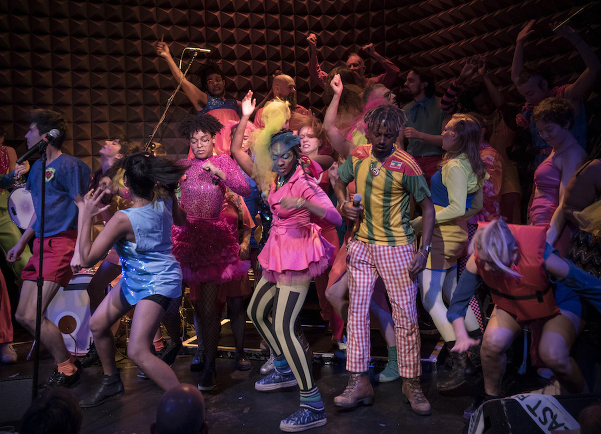 A photograph of the Stop Shopping Choir, a group of young people in colourful clothes are mid-dance.