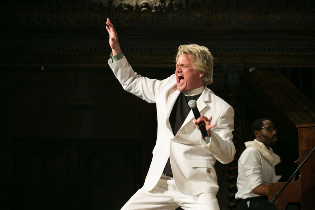 A man dressed in a white suit and a priest's collar shouts into a mic and raises one arm excitedly.