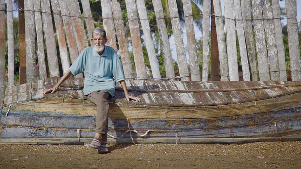 A man with a white beard sits on an upturned boat with a rickety fence behind him.