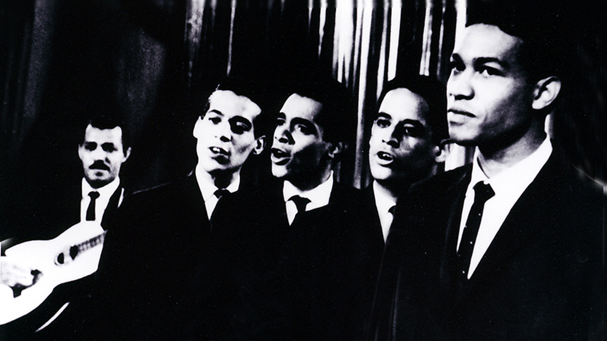 A black and white image of five men in suits and ties, the one on the left holding a guitar.